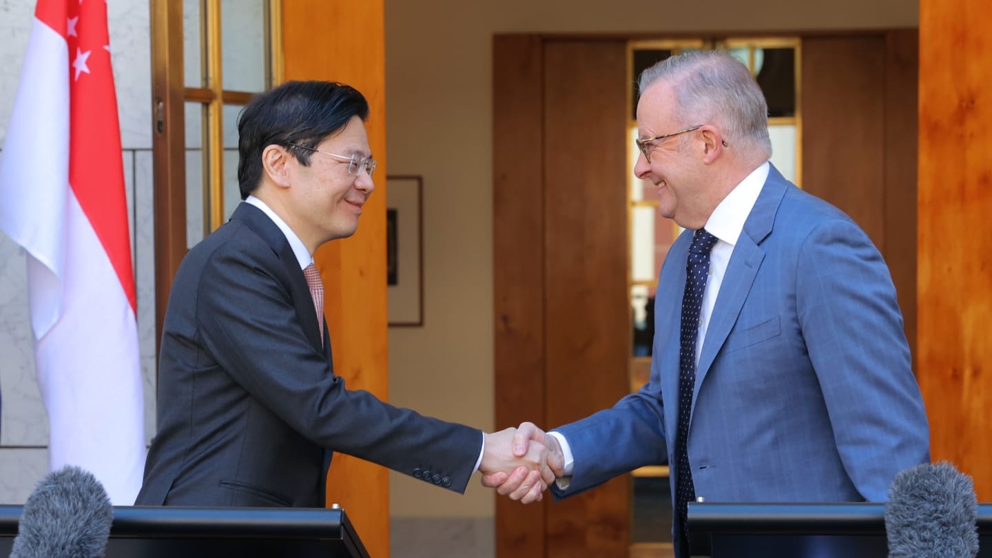 Two leaders in suits shake hands at podiums outside; Singapore flag and microphones visible.